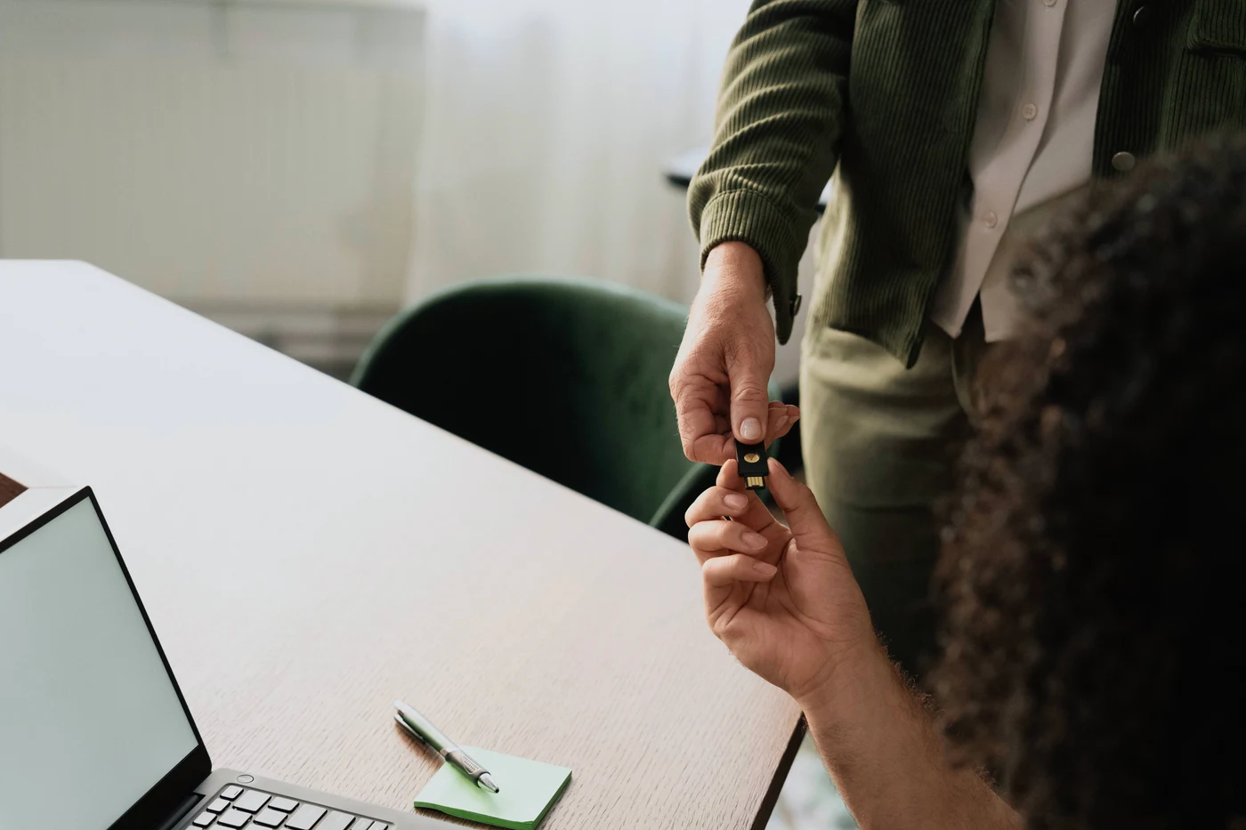 Ruka pruža sigurnosni ključ YubiKey s USB-A priključkom kolegi za uredskim stolom s otvorenim prijenosnikom